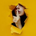 mulher saindo de um rasgo aberto em uma parede amarela, com o dedo indicador sobre os lábios pedindo silêncio