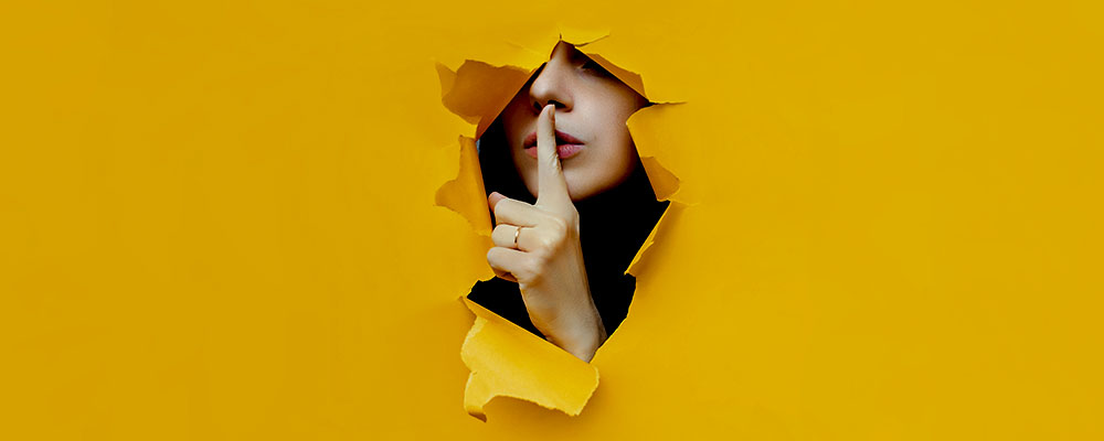 mulher saindo de um rasgo aberto em uma parede amarela, com o dedo indicador sobre os lábios pedindo silêncio