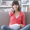 mulher sentada no sofá com uma bacia de pipoca, apontando o controle para a TV, para assistir séries brasileiras na Netflix