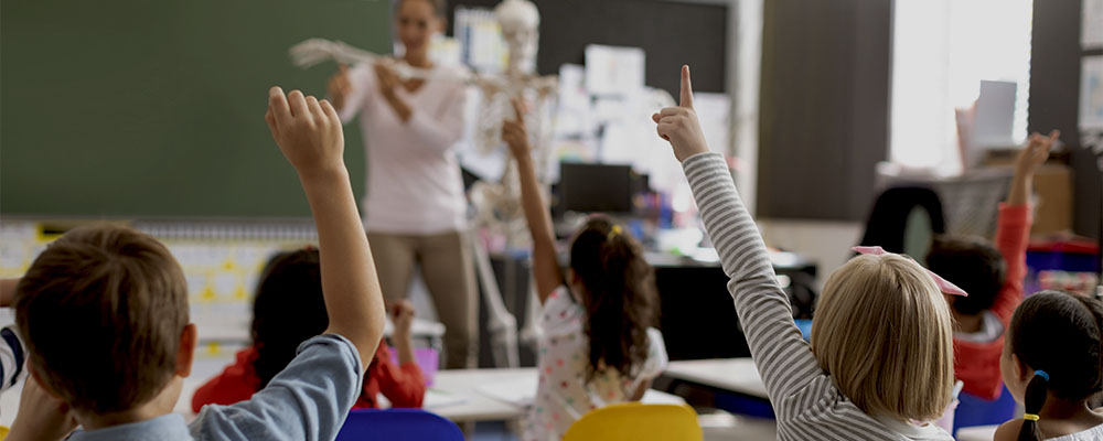 Crianças em uma sala de aula levantando as mãos, mostrando que são alunos participativos e estão tendo uma educação de qualidade