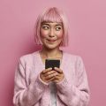 garota de cabelo e roupa cor de rosa usando um celular e olhando para o lado dando um sorriso torto