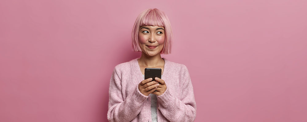 garota de cabelo e roupa cor de rosa usando um celular e olhando para o lado dando um sorriso torto