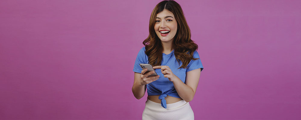 mulher sorrindo e usando celular sobre um fundo rosa
