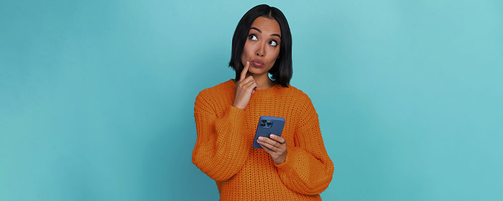 mulher segurando um celular com o dedo no queixo indicando dúvida