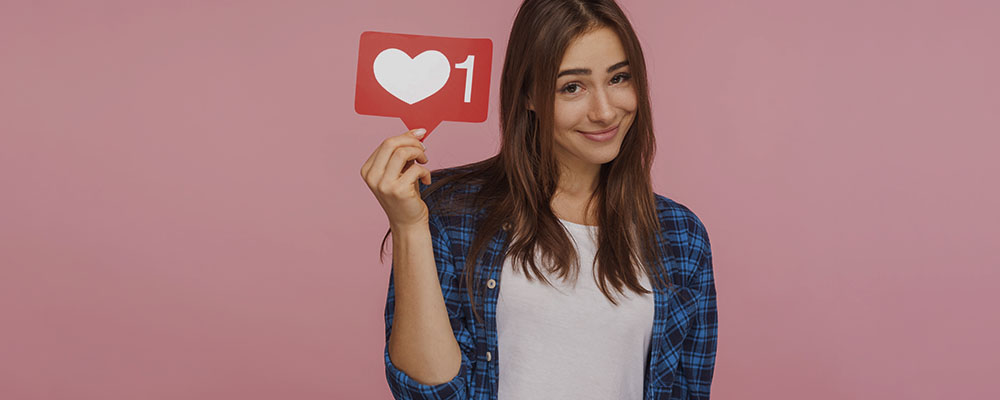 garota segurando uma plaquinha com símbolo de coração e o número 1 ao lado, representando os likes de redes sociais