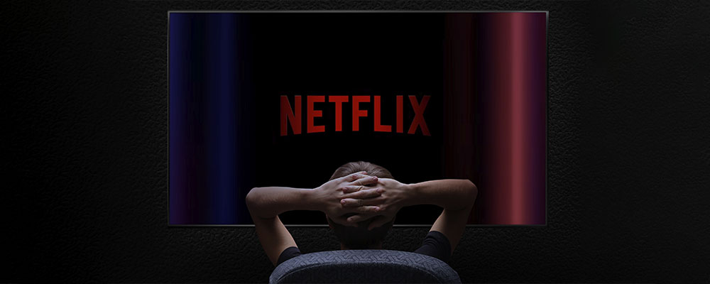 homem sentado com as mãos atrás da cabeça assistindo Netflix na televisão