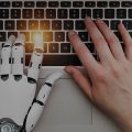 mão de um robô ao lado da mão de uma mulher digitando no teclado de um computador