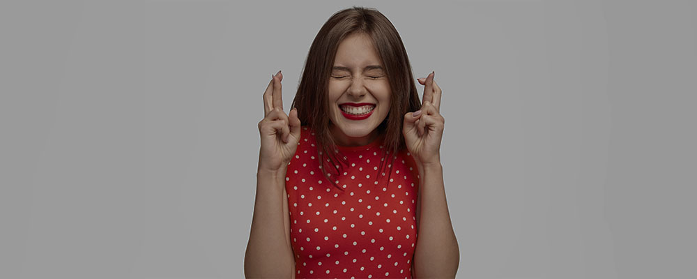 mulher sorrindo com os olhos fechados e fazendo figas com as duas mãos, esperando ter sorte em um sorteio