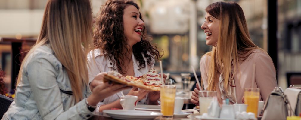 Amigas sorrindo enquanto comem pizza no exterior de um restaurante.