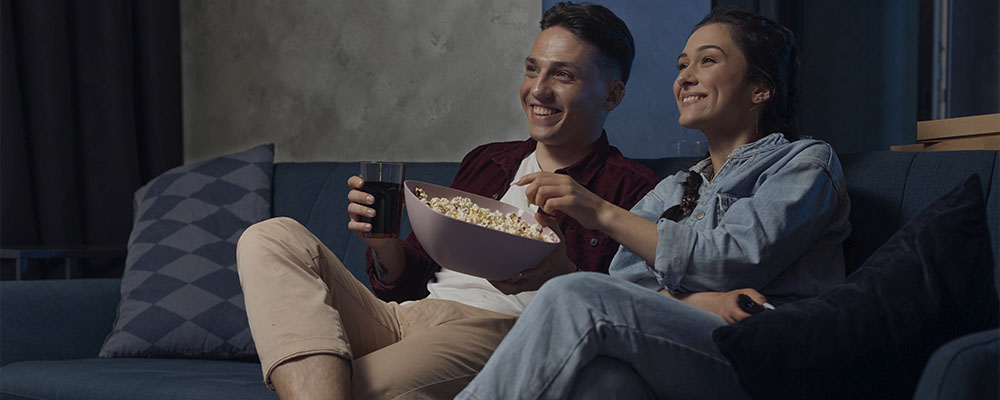 Casal assistindo séries da Netflix sentados em um sofá comendo pipoca.