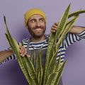 Homem atrás de uma planta, abrindo as folhagens com as mãos e sorrindo.