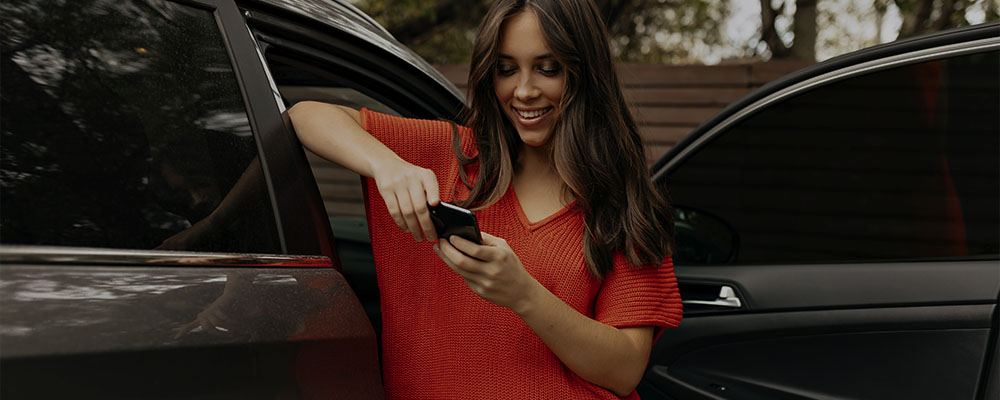 Mulher encostada na porta aberta de um carro enquanto usa seu celular para organizar a revisão do carro.