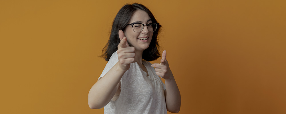 Garota sorrindo e piscando um olho enquanto aponta para a câmera de um jeito divertido, como quem está dando uma dica.