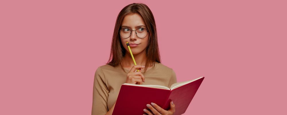 Garota pensando na redação do ENEM segurando um caderno e olhando para cima com um lápis apoiado no queixo.