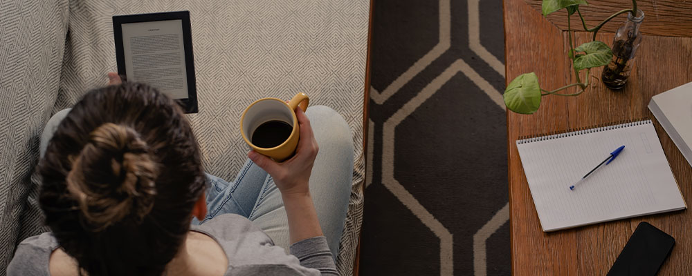 Mulher lendo no Kindle no sofá enquanto toma uma xícara de café.