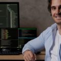 Homem que trabalha com TI sentado diante de sua mesa com 3 monitores exibindo linhas de código de programação.