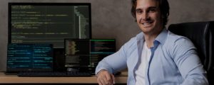 Homem que trabalha com TI sentado diante de sua mesa com 3 monitores exibindo linhas de código de programação.