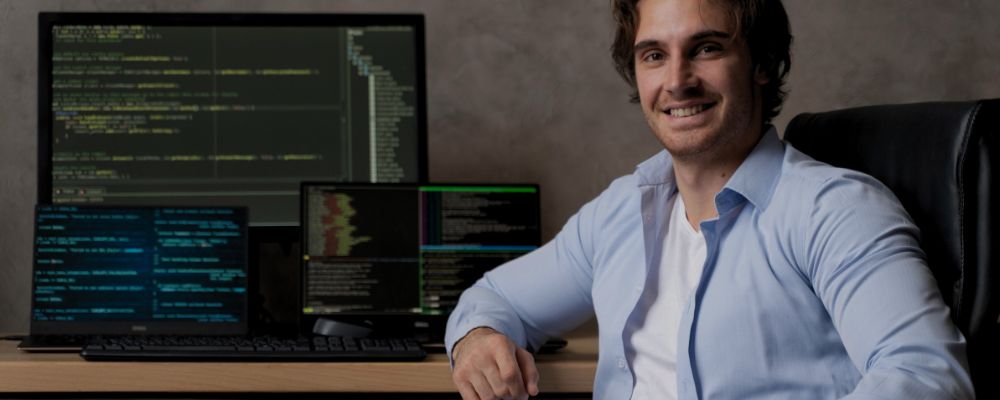Homem que trabalha com TI sentado diante de sua mesa com 3 monitores exibindo linhas de código de programação.