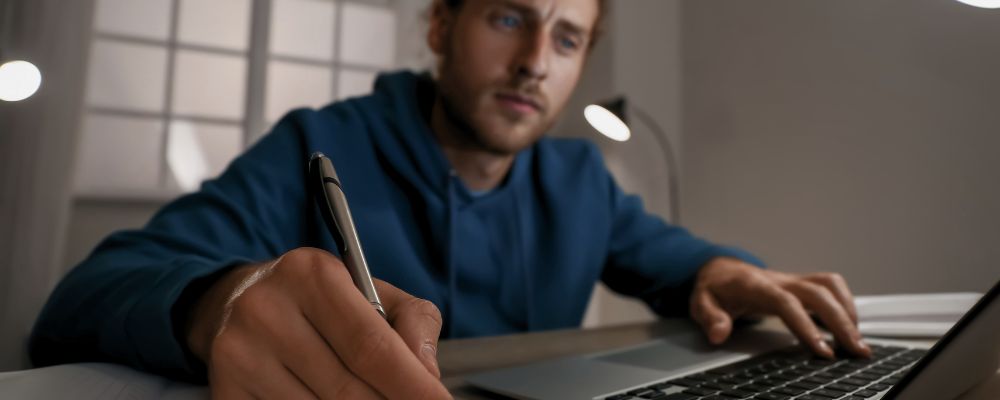 Homem assistindo aula online em um notebook e fazendo anotações em um caderno.