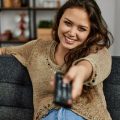 Mulher sentada no sofá apontando o controle para a TV para assistir os lançamentos da Netflix de junho de 2024.