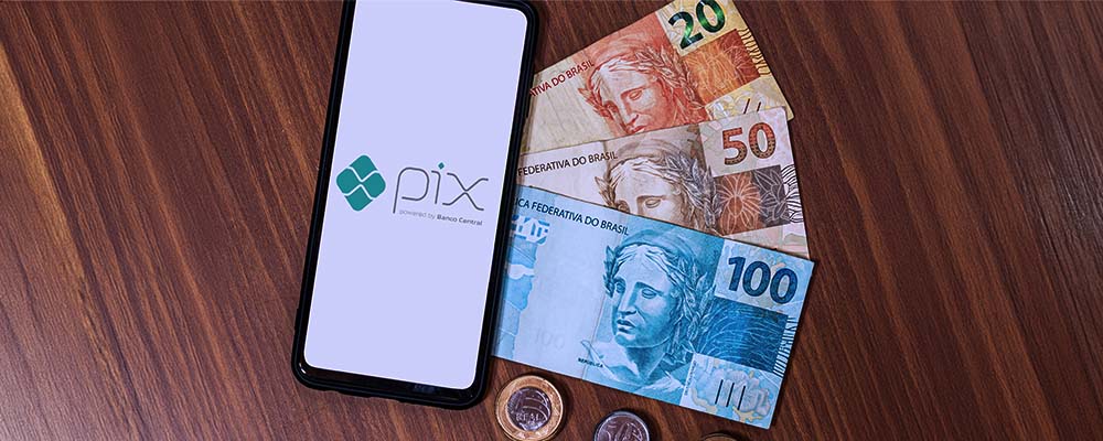 Celular com o logo do Pix ao lado de cédulas e moedas de real.