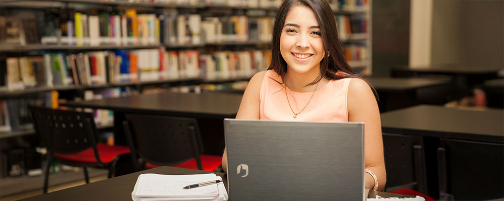 Mulher estudando na biblioteca da faculdade com um notebook Positivo.