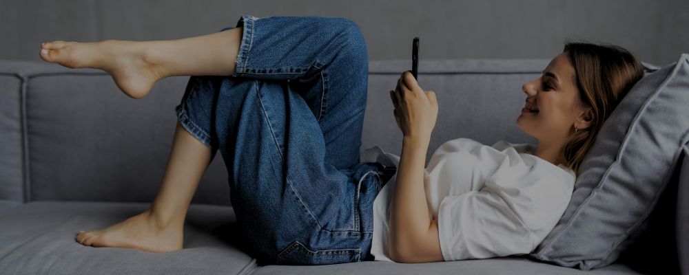 Mulher deitada no sofá controlando sua casa por meio de assistentes virtuais.