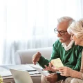 Casal de idosos, um homem e uma mulher, sentados confortavelmente em casa, analisando documentos e notas de dinheiro. O homem, com óculos e suéter verde, segura papéis, enquanto a mulher, com colar de pérolas, segura cédulas. Eles estão focados em planejamento financeiro, orçamento doméstico ou finanças da aposentadoria, com um laptop visível na mesa de centro.