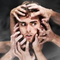 Rosto de um homem jovem com cabelo castanho claro e olhos azuis arregalados, olhando para o lado com uma expressão de surpresa ou angústia. Múltiplas mãos de diferentes pessoas tocam e seguram sua cabeça e rosto, simbolizando pressão, estresse, confusão ou sobrecarga mental. O fundo é escuro e desfocado, destacando o drama da cena.