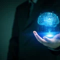 Mão de um profissional de negócios segurando um holograma azul brilhante de um cérebro digital com o ícone 'AI' (Inteligência Artificial) no centro, simbolizando inovação tecnológica, aprendizado de máquina e o futuro da IA.