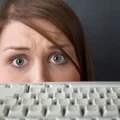 Close-up dos olhos de uma mulher com cabelo castanho e expressão de surpresa ou apreensão, olhando por cima de um teclado de computador branco e desfocado. A imagem sugere preocupação com segurança digital, privacidade online ou um erro no trabalho.