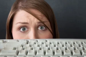 Close-up dos olhos de uma mulher com cabelo castanho e expressão de surpresa ou apreensão, olhando por cima de um teclado de computador branco e desfocado. A imagem sugere preocupação com segurança digital, privacidade online ou um erro no trabalho.