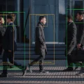 Vista lateral de diversas pessoas, incluindo profissionais de negócios, caminhando em uma calçada urbana. Caixas de detecção e reconhecimento facial de inteligência artificial sobre as figuras indicam monitoramento tecnológico. Ao fundo, um edifício moderno de vidro reflete a luz do sol.