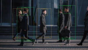 Vista lateral de diversas pessoas, incluindo profissionais de negócios, caminhando em uma calçada urbana. Caixas de detecção e reconhecimento facial de inteligência artificial sobre as figuras indicam monitoramento tecnológico. Ao fundo, um edifício moderno de vidro reflete a luz do sol.