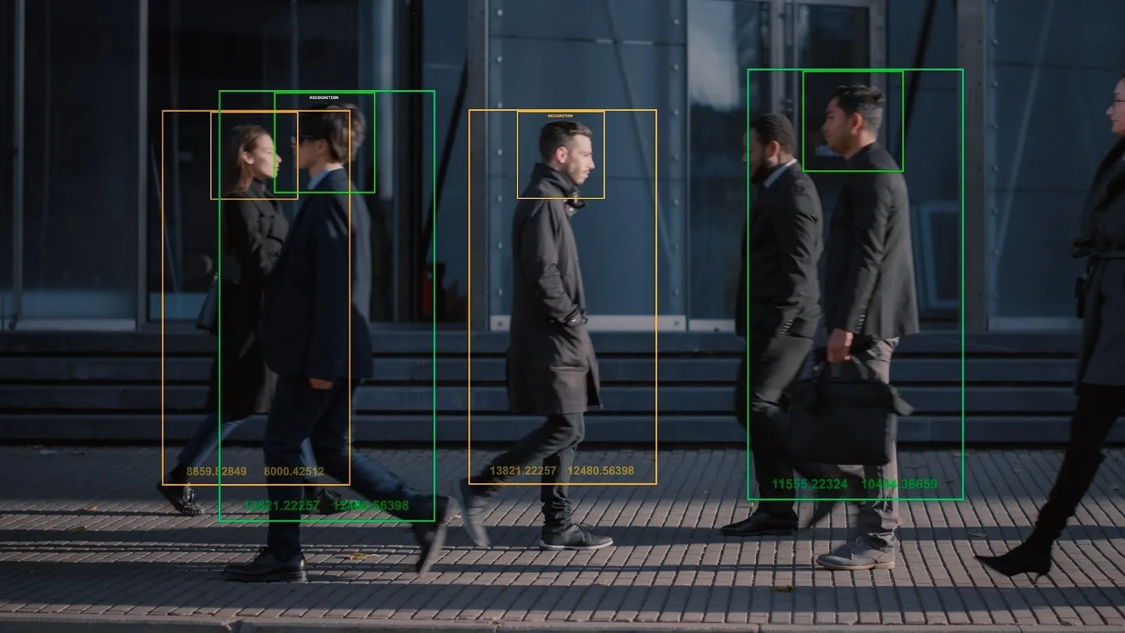 Vista lateral de diversas pessoas, incluindo profissionais de negócios, caminhando em uma calçada urbana. Caixas de detecção e reconhecimento facial de inteligência artificial sobre as figuras indicam monitoramento tecnológico. Ao fundo, um edifício moderno de vidro reflete a luz do sol.