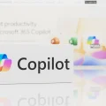 Logo do Microsoft Copilot em um bloco branco com o texto 'Copilot' e o símbolo colorido, refletido em uma superfície. Ao fundo, uma tela desfocada exibe o slogan 'Reinvente a produtividade com Microsoft 365 Copilot' e ícones de aplicativos como Excel, Word e PowerPoint, destacando a integração da inteligência artificial para otimização de trabalho e colaboração.