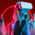 Mulher loira com óculos de realidade virtual (VR) brancos, imersa em um ambiente futurista com iluminação neon vermelha e azul. Ela está com as mãos levantadas, interagindo com o mundo virtual, vestindo uma jaqueta transparente. A imagem evoca tecnologia, inovação e metaverso.