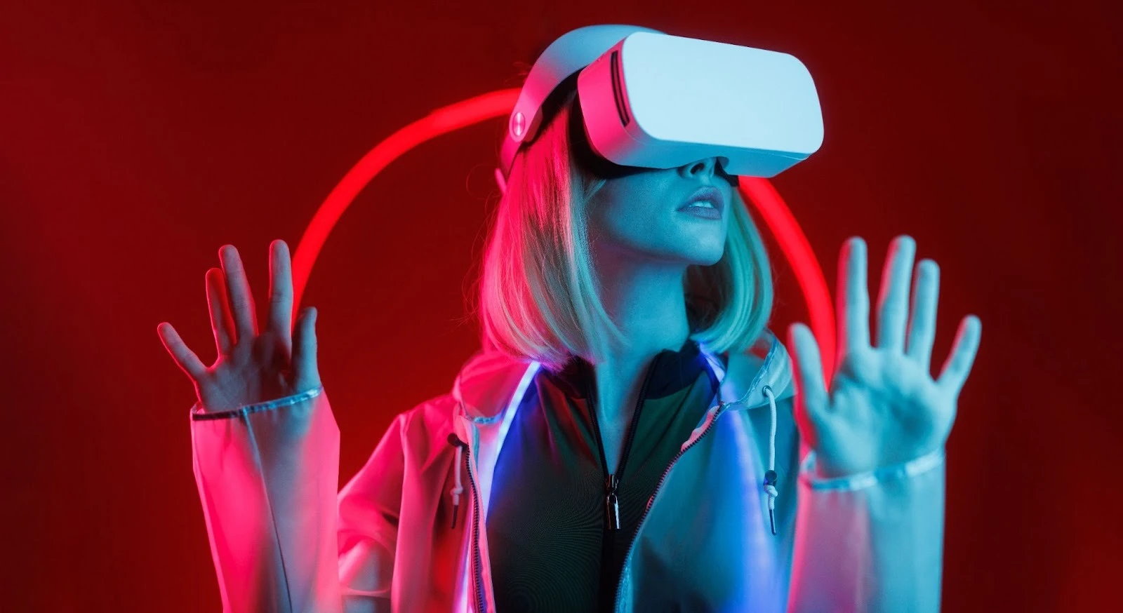 Mulher loira com óculos de realidade virtual (VR) brancos, imersa em um ambiente futurista com iluminação neon vermelha e azul. Ela está com as mãos levantadas, interagindo com o mundo virtual, vestindo uma jaqueta transparente. A imagem evoca tecnologia, inovação e metaverso.