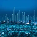 Paisagem urbana noturna com prédios e arranha-céus iluminados, sobreposta por linhas de luz verticais e pontos brilhantes, simbolizando rede de dados, conectividade tecnológica e comunicação global em uma cidade inteligente.