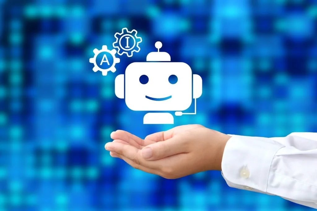 Mão de uma pessoa em camisa branca segurando um ícone flutuante de um robô chatbot branco sorridente com fones de ouvido, simbolizando inteligência artificial (IA) e automação. Acima do robô, ícones de engrenagens com as letras 'A' e 'I' destacam a tecnologia de IA. O fundo digital azul desfocado reforça o tema tecnológico. Perfeito para ilustrar conceitos de atendimento ao cliente automatizado, suporte digital e inovação tecnológica.