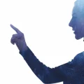 Silhueta de uma mulher em perfil apontando com o dedo indicador para uma superfície ou interface virtual invisível. A imagem tem um efeito de dupla exposição com partículas digitais ou estrelas em tons de azul e branco, simbolizando tecnologia, inovação, realidade aumentada ou interação futurista em um fundo branco.
