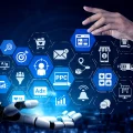 Uma mão humana e uma mão robótica interagem com uma projeção holográfica azul de ícones de marketing digital e e-commerce, incluindo PPC, Ads, análise de dados, segmentação de público-alvo, carrinho de compras, email marketing, A/B testing e logística de entrega. O fundo é um ambiente digital escuro com linhas de rede e luzes borradas, simbolizando a integração de inteligência artificial e automação nas estratégias de marketing online e vendas digitais.