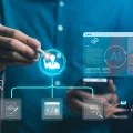 Mão de um profissional usando uma caneta stylus para interagir com uma interface digital holográfica que representa um sistema de inteligência artificial (IA) e gestão de dados. A tela exibe um ícone central de um especialista, gráficos de análise de dados (barras, pizza, linhas), documentos, código, e fluxos de trabalho complexos interligando perfis de usuário, dados e comunicação digital. A imagem simboliza consultoria tecnológica, otimização de processos e business intelligence com IA.
